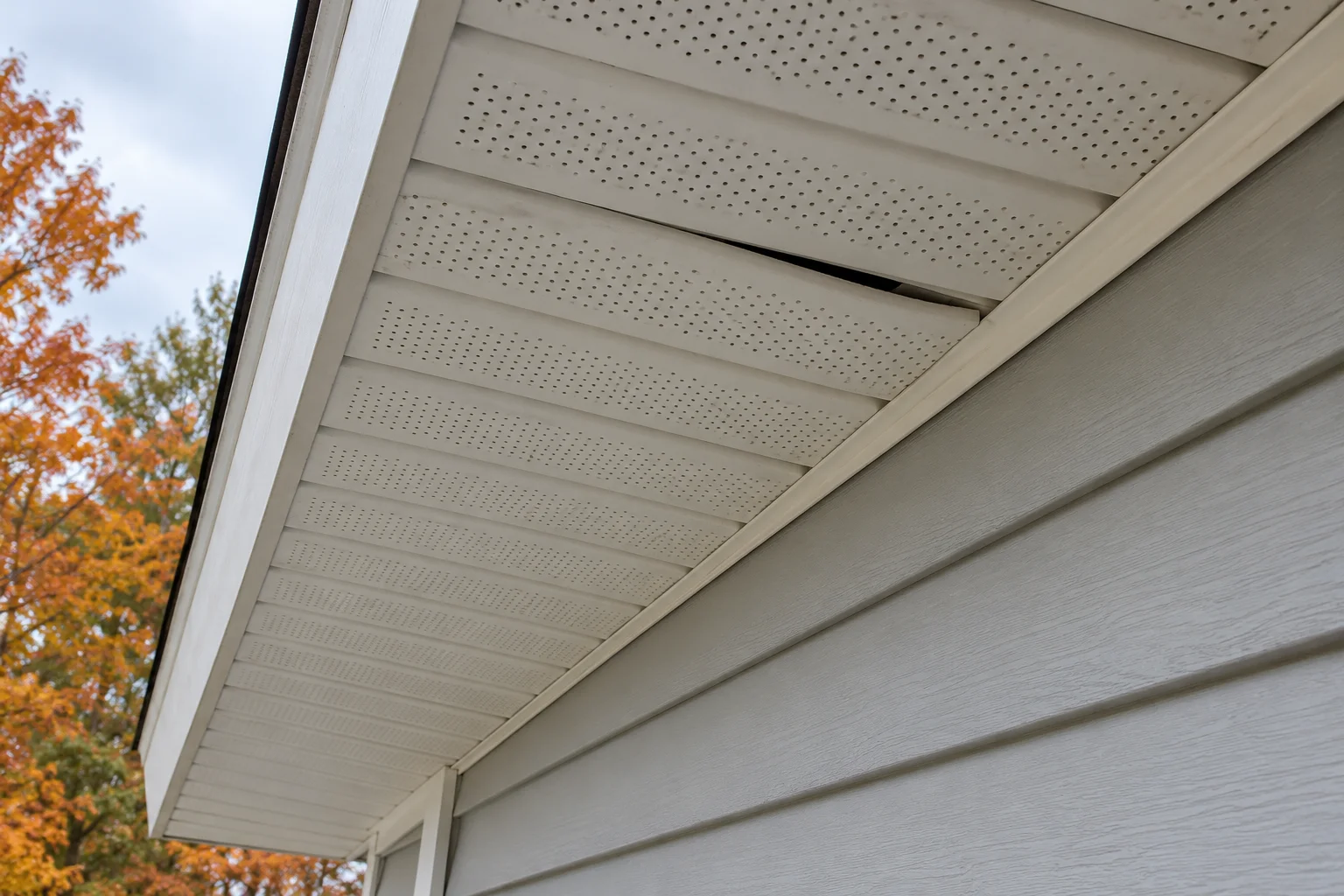 White perforated soffit panels beneath a roof overhang, with a small loose panel edge above gray siding